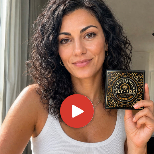 A woman with curly dark hair holds up a black decorative box labeled 'Sly-Fox' and 'Hagover Patch' while wearing a white ribbed tank top. A red play button overlay is centered on the image.
